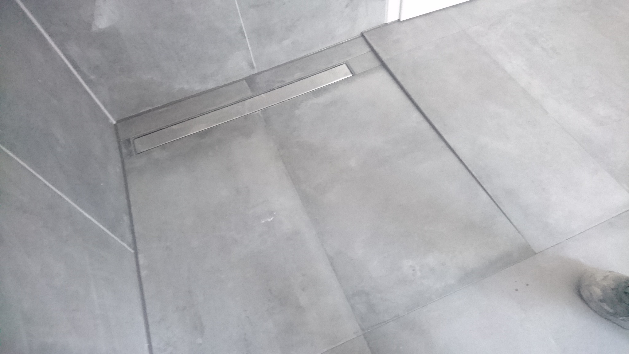 Szare płytki podłogowe i ścienne w łazience z odpływem liniowym, widoczne fugi i fragment buta w prawym dolnym rogu.