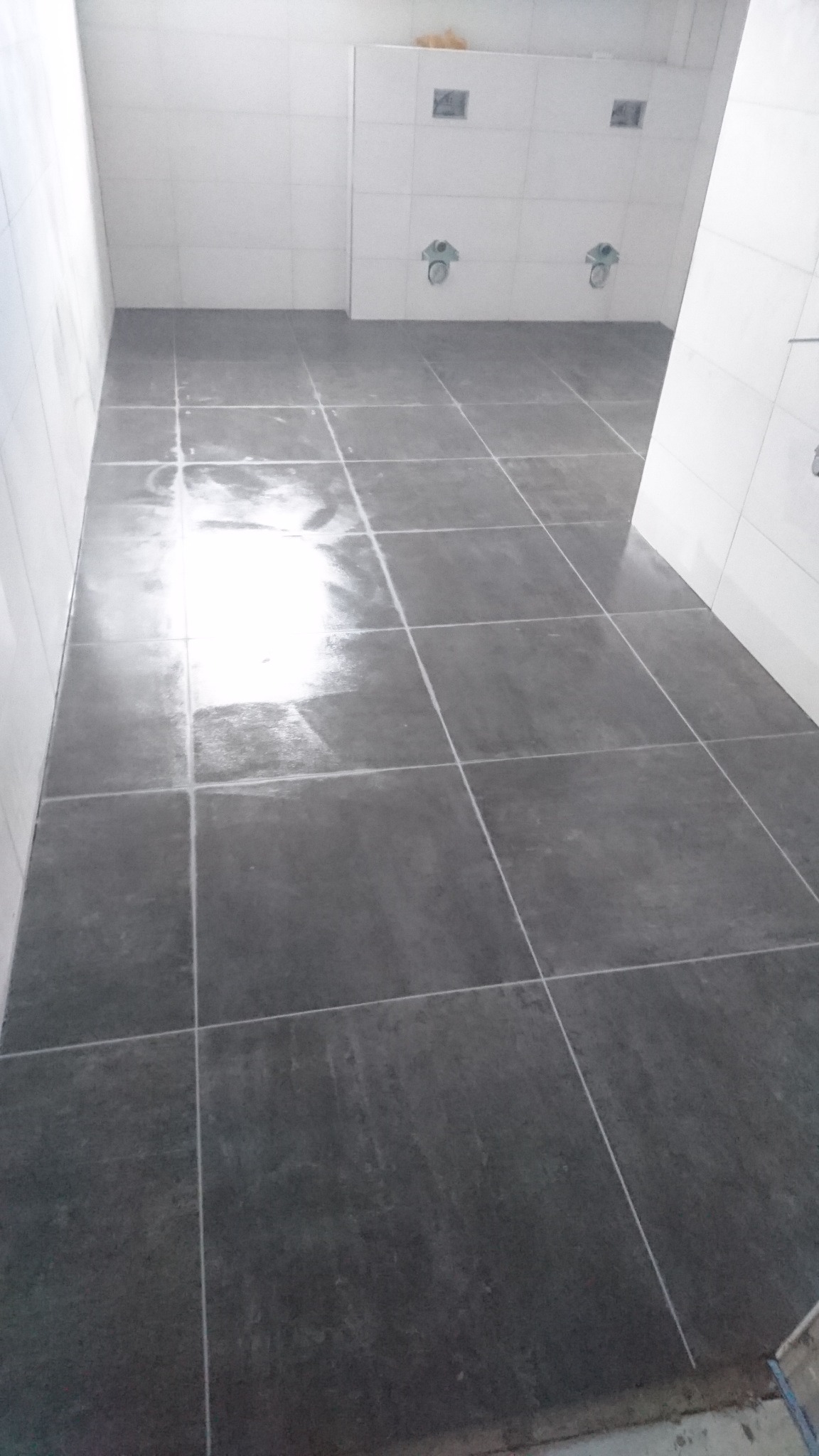 Świeżo położona, ciemnoszara, błyszcząca podłoga z płytek w łazience z białymi, częściowo wykończonymi ścianami i instalacjami sanitarnymi.