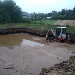 Wykop pod fundamenty zalany wodą, z widocznym minikoparką i zagęszczarkami na placu budowy, w tle dom jednorodzinny.