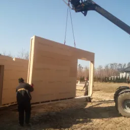 Montaż prefabrykowanej ściany z otworem okiennym za pomocą dźwigu teleskopowego na placu budowy, widoczni dwaj pracownicy w odzieży roboczej.