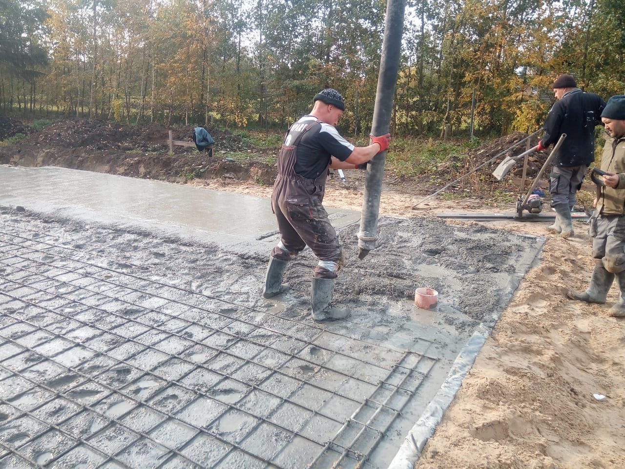 Wylewanie betonu na siatkę zbrojeniową pod fundament domu, pracownicy w kombinezonach i kaloszach, jeden z nich trzyma rurę do betonu, w tle drzewa.