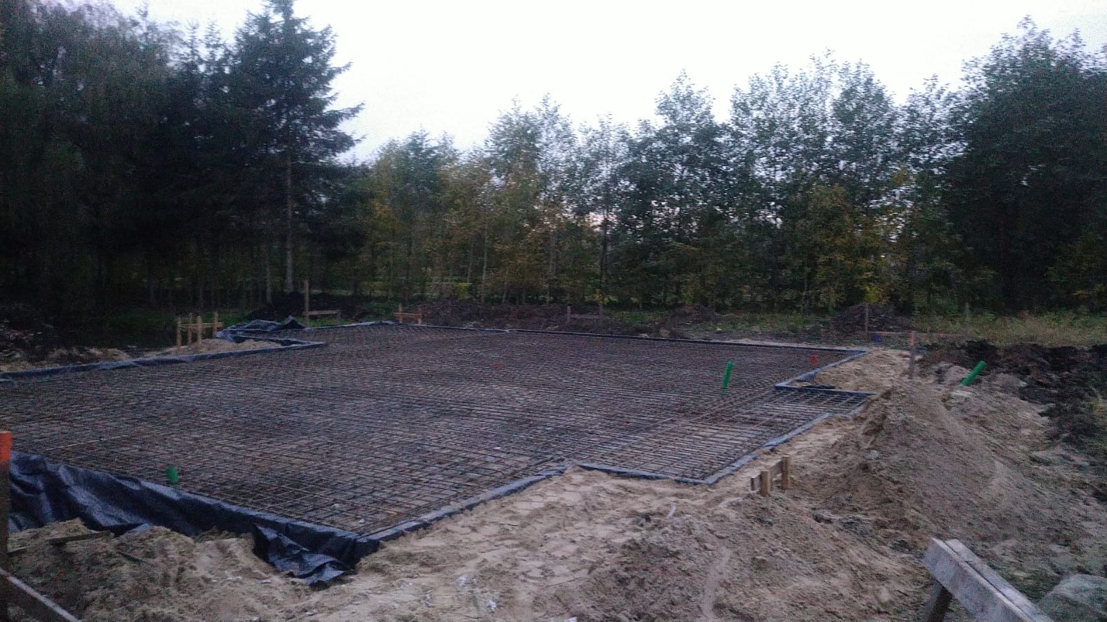 Przygotowanie zbrojenia płyty fundamentowej pod budowę, widoczne siatki zbrojeniowe, folia izolacyjna, pionowe rury i drewniane szalunki, w tle las.
