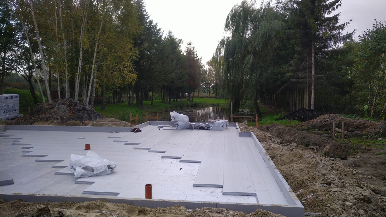 Przygotowanie fundamentów pod budowę domu: izolacja termiczna w formie płyt styropianowych ułożona na betonowej wylewce, widok na działkę z drzewami i małym stawem w tle.