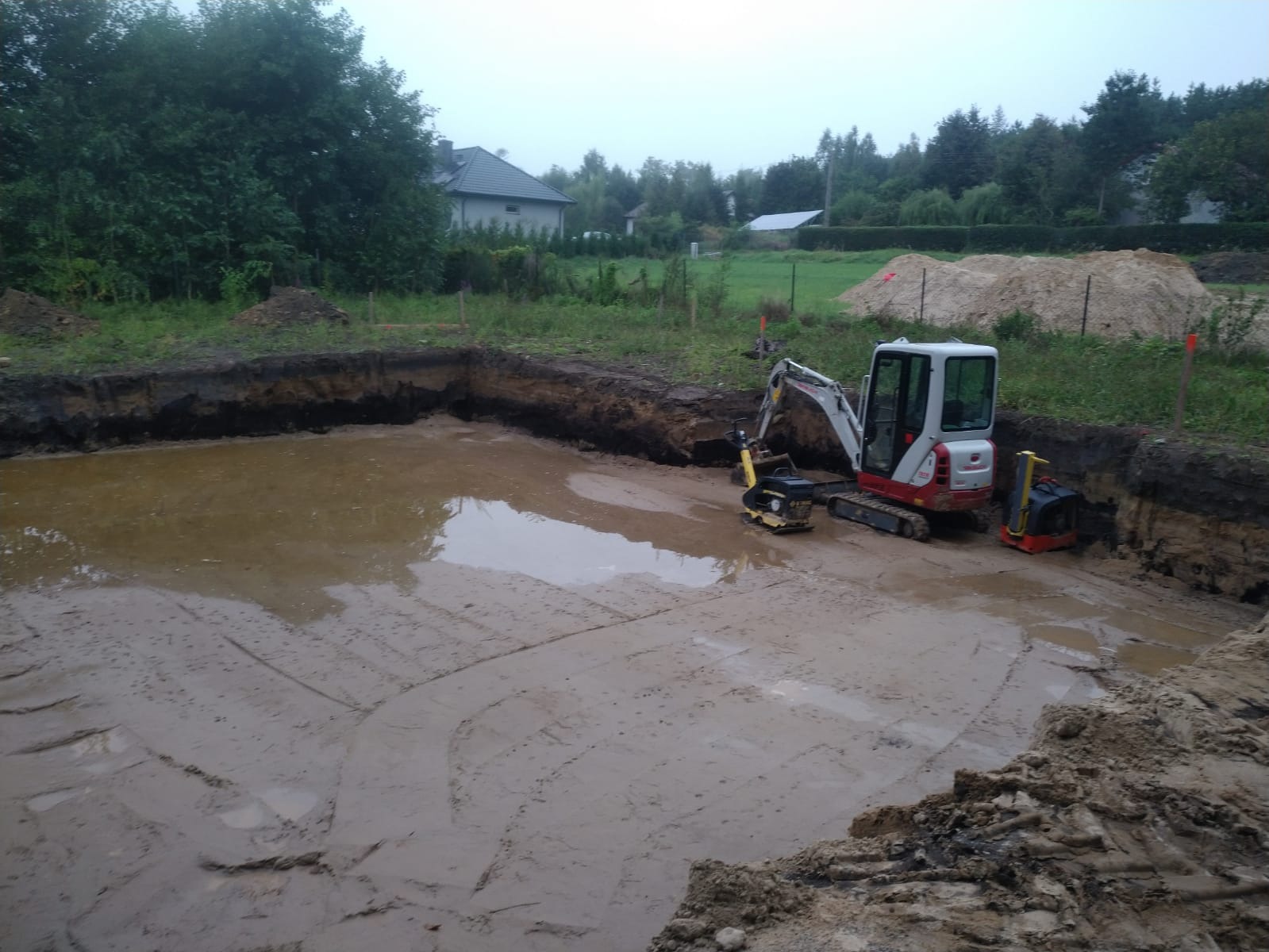 Wykop pod fundamenty zalany wodą, z widocznym minikoparką i zagęszczarkami na placu budowy, w tle dom jednorodzinny.