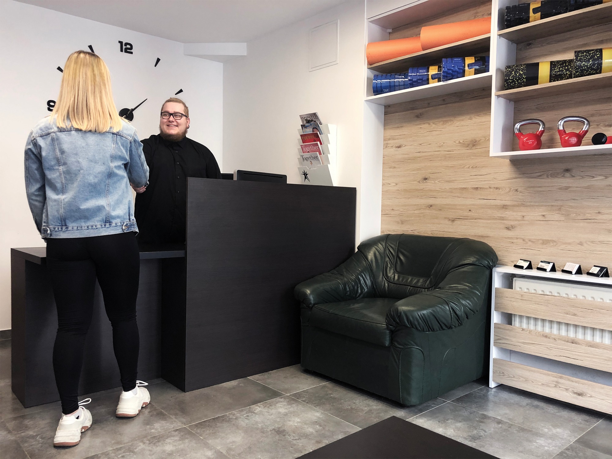 Recepcja w centrum rehabilitacji: blondynka w jeansowej kurtce podaje rękę uśmiechniętemu mężczyźnie za czarną ladą, w tle zegar ścienny, półki z matami do ćwiczeń, kettlebells i zielony skórzany...