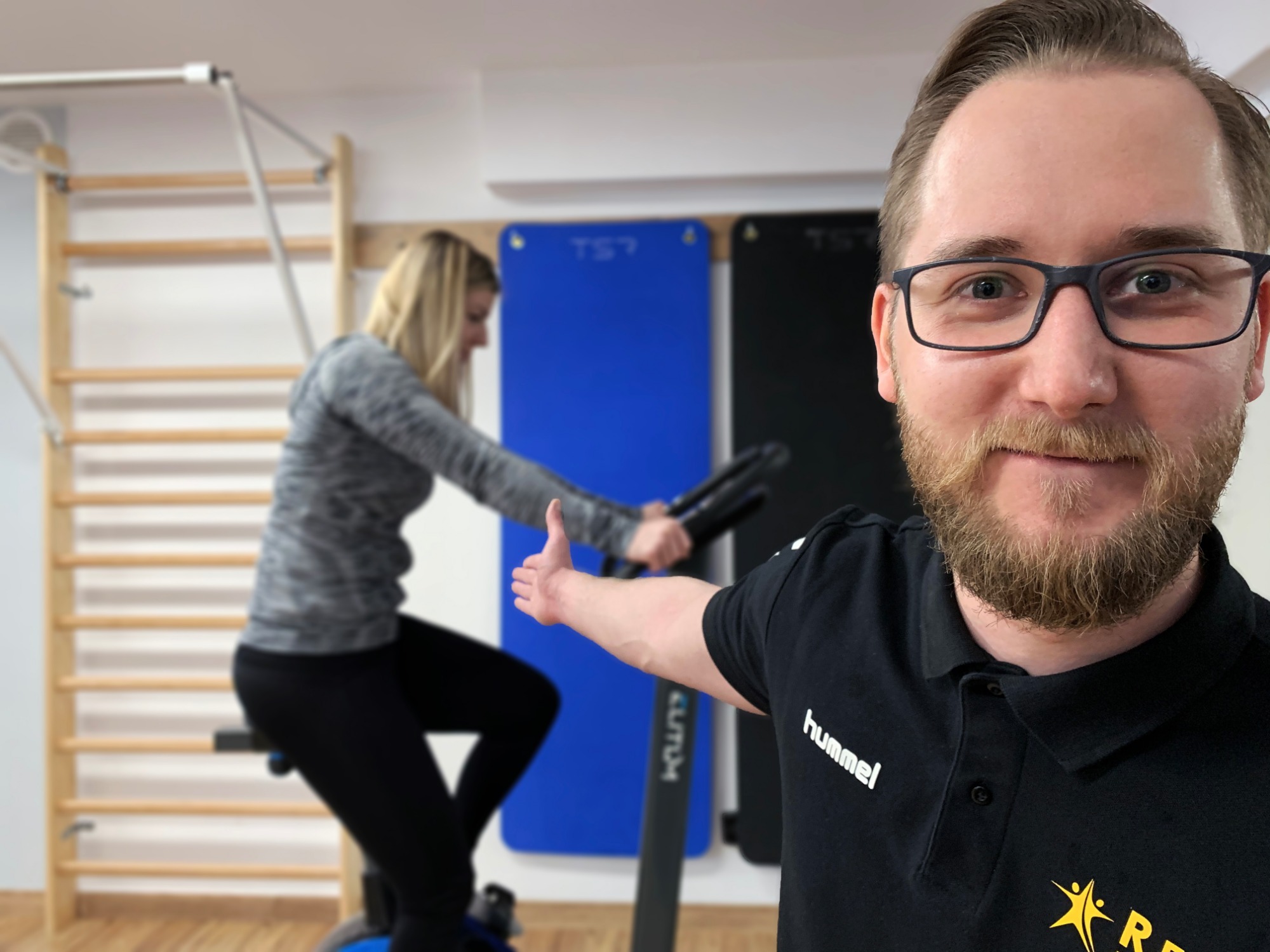 Uśmiechnięty mężczyzna w okularach i czarnej koszulce polo prezentuje ćwiczącą kobietę na rowerze stacjonarnym w tle, drabinki gimnastyczne i tablice edukacyjne w gabinecie.