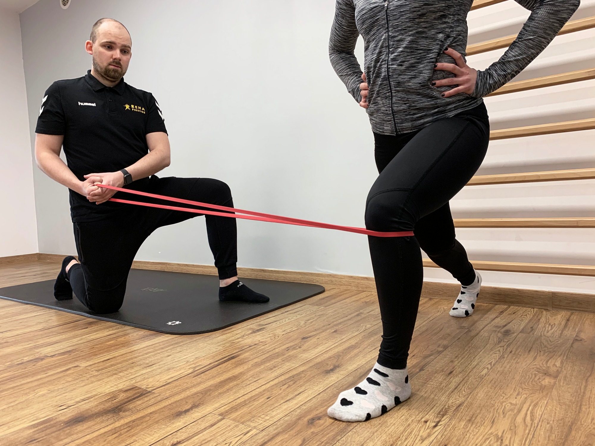 Osoba wykonuje wykrok z gumą oporową pod nadzorem trenera, widoczna mata do ćwiczeń i drabinka gimnastyczna.