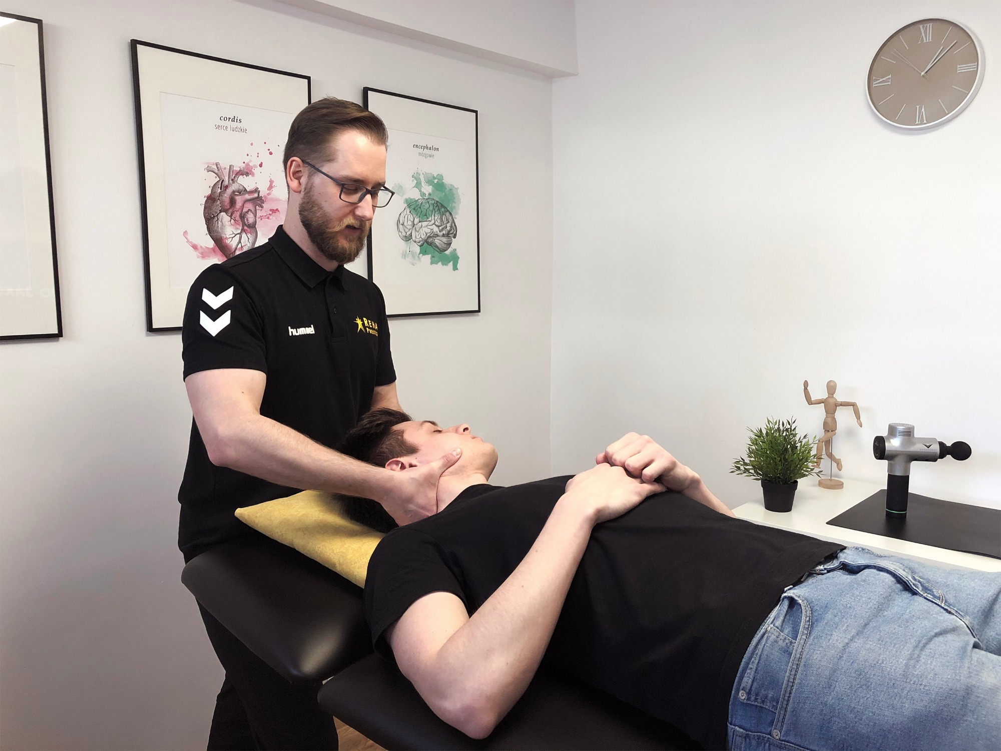 Mężczyzna w okularach w czarnej koszulce z logo firmy wykonuje terapię manualną szyi pacjenta leżącego na kozetce z żółtą poduszką. W tle białe ściany z obrazami anatomicznymi i zegarem.