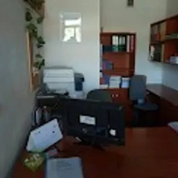 Biuro z drewnianym biurkiem, komputerem i kalendarzem na pierwszym planie, w tle regał z książkami i segregatorami. Na ścianie wisi lustro.