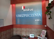 Recepcja biura ubezpieczeniowego Brokus z podświetlonym logo i napisem UBEZPIECZENIA od 1995 r. na tle ściany w kolorze srebrnym, otoczona drewnianą ramą.