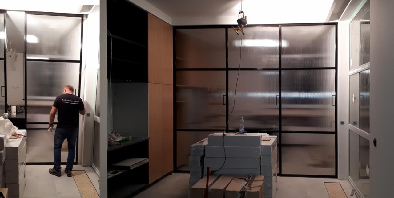 Montaż ścianki działowej z czarnych profili i szkła ryflowanego, widoczny pracownik w koszulce firmowej i przygotowane elementy konstrukcyjne.