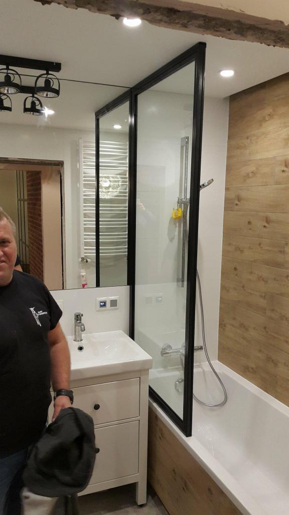 Nowoczesna łazienka z kabiną prysznicową w czarnej ramie, umywalką na szafce i drewnopodobną okładziną ściany, widoczny fragment osoby w czarnej koszulce.