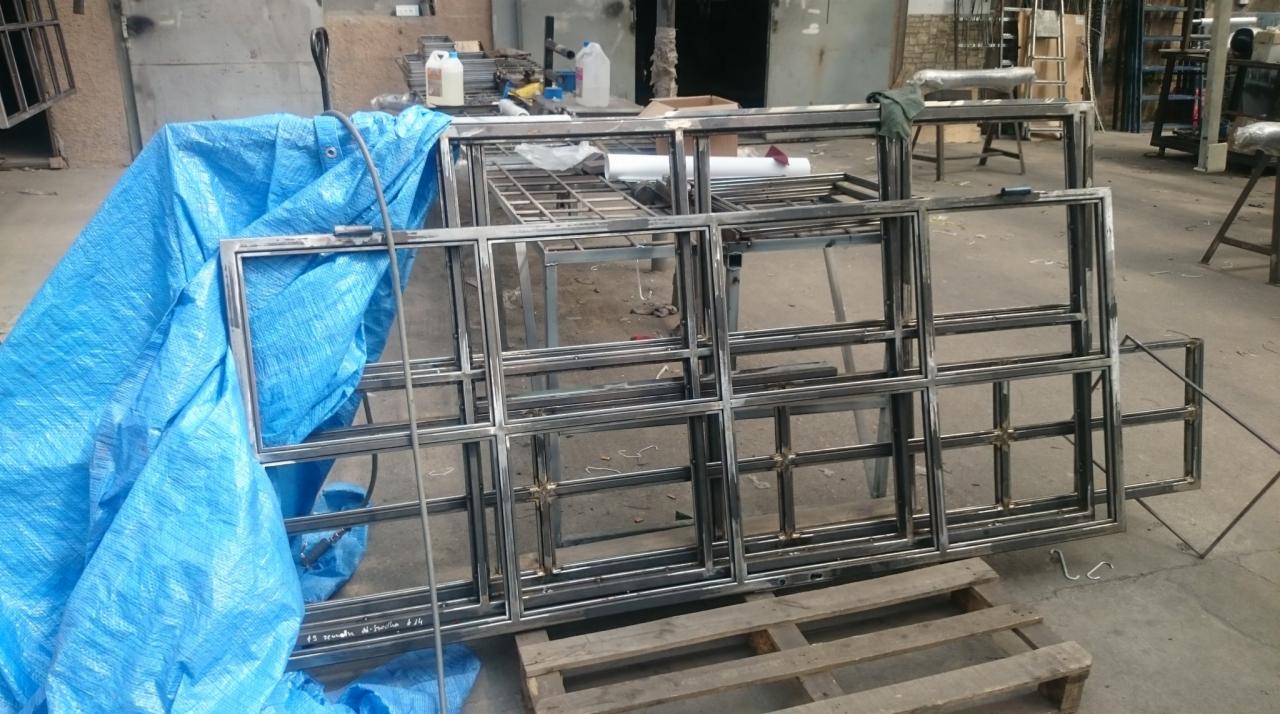 Szkielety metalowych konstrukcji z profili kwadratowych, częściowo przykryte niebieską plandeką, ułożone na drewnianej palecie w warsztacie.