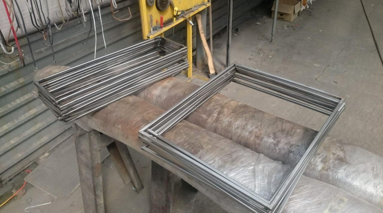 Stos metalowych, kwadratowych ram spawanych z rurek, ułożonych na zabezpieczonych folią rurach w warsztacie z narzędziami w tle.