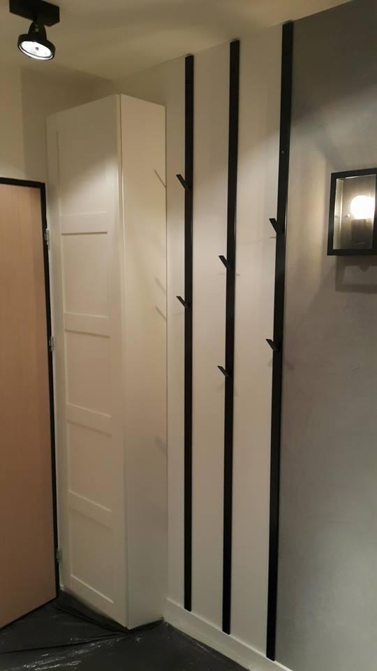 Wnętrze przedpokoju z białą szafką i trzema pionowymi wieszakami ściennymi z czarnymi elementami, oświetlone punktowym światłem.