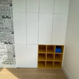 Biała szafa wnękowa z żółtą wnęką na zabawki, z pojedynczym niebieskim klockiem Lego, na tle ściany w tapetę z motywem domków.