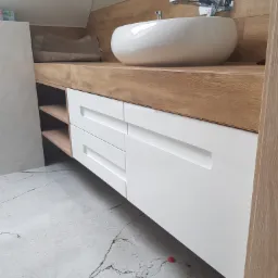 Nowoczesna łazienka na poddaszu: biała umywalka nablatowa i szafka z drewnianym blatem w jasnym odcieniu drewna, szuflady z prostokątnymi uchwytami, widoczny fragment podłogi z szarego marmuru...