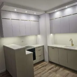 Nowoczesna kuchnia w odcieniach szarości z drewnianą podłogą, oświetleniem LED podszafkowym i złotymi akcentami na górnych szafkach.