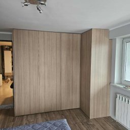Meble na Wymiar DOMEB Łukasz Pamuła - Jasna szafa wnękowa z drewnianym wykończeniem, dopasowana do rogu pokoju. Widoczny fragment łóżka z szarą narzutą i pies leżący w tle.