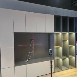 Meble na Wymiar DOMEB Łukasz Pamuła - Montaż szafy wnękowej z użyciem poziomicy laserowej. Widoczne półki i zamknięte szafki w odcieniach szarości. Podłoga drewniana.