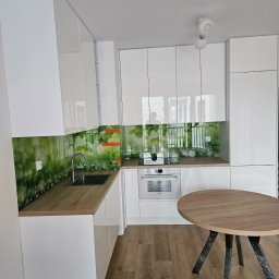 Meble na Wymiar DOMEB Łukasz Pamuła - Biała kuchnia z blatem drewnopodobnym i szklanym panelem z motywem roślinnym. Okrągły stół z drewnianym blatem na metalowych nogach. Nowoczesne oświetlenie sufitowe.