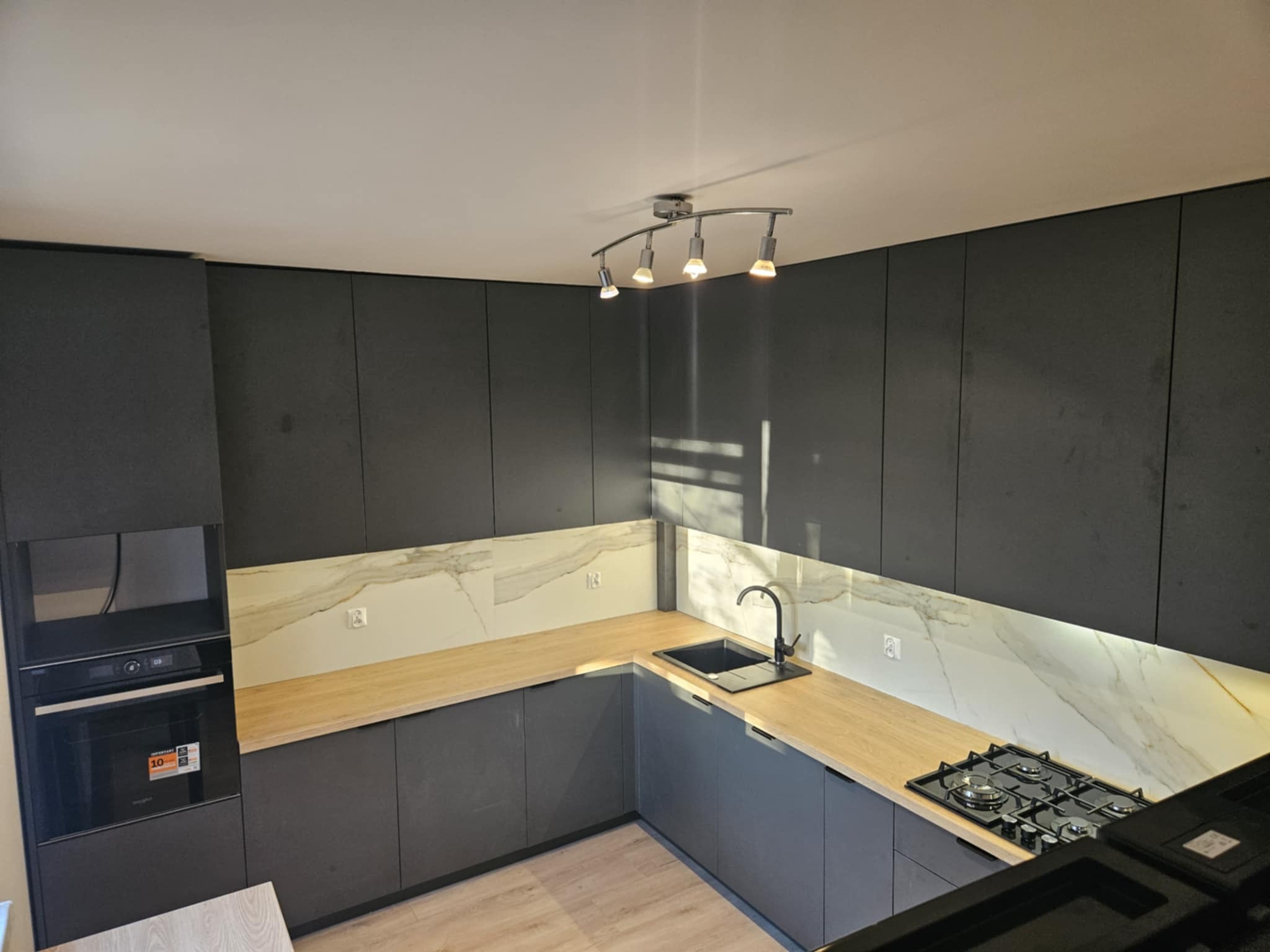 Nowoczesna kuchnia z ciemnymi, matowymi frontami szafek, drewnianym blatem, marmurowym panelem nad blatem i oświetleniem punktowym.
