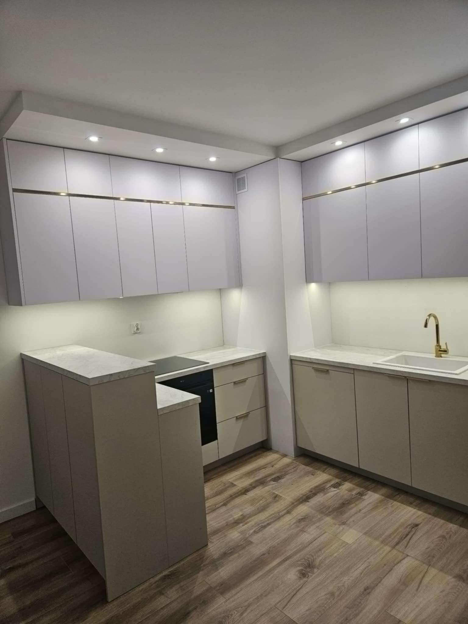 Nowoczesna kuchnia w odcieniach szarości z drewnianą podłogą, oświetleniem LED podszafkowym i złotymi akcentami na górnych szafkach.