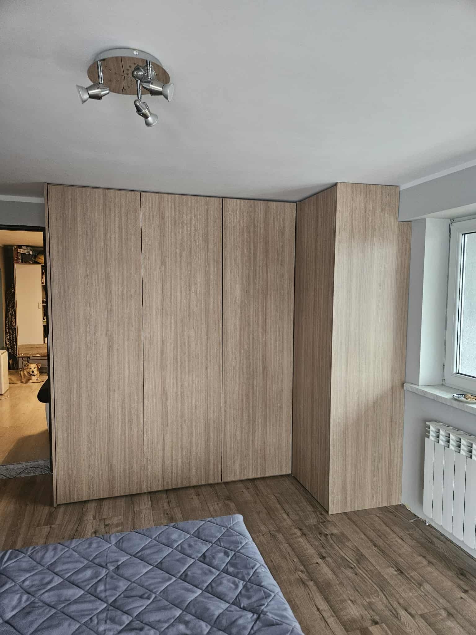 Jasna szafa wnękowa z drewnianym wykończeniem, dopasowana do rogu pokoju. Widoczny fragment łóżka z szarą narzutą i pies leżący w tle.