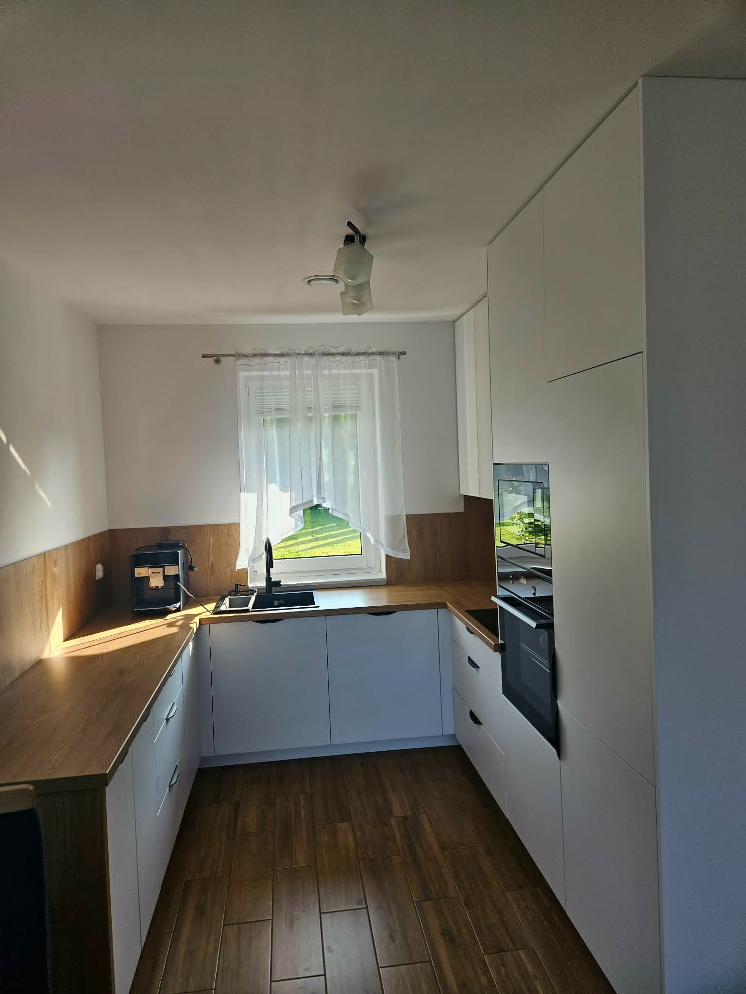 Jasna kuchnia w kształcie litery U z drewnianym blatem i białymi frontami. Widoczne okno z firanką i ekspres do kawy. Podłoga wyłożona panelami imitującymi drewno.