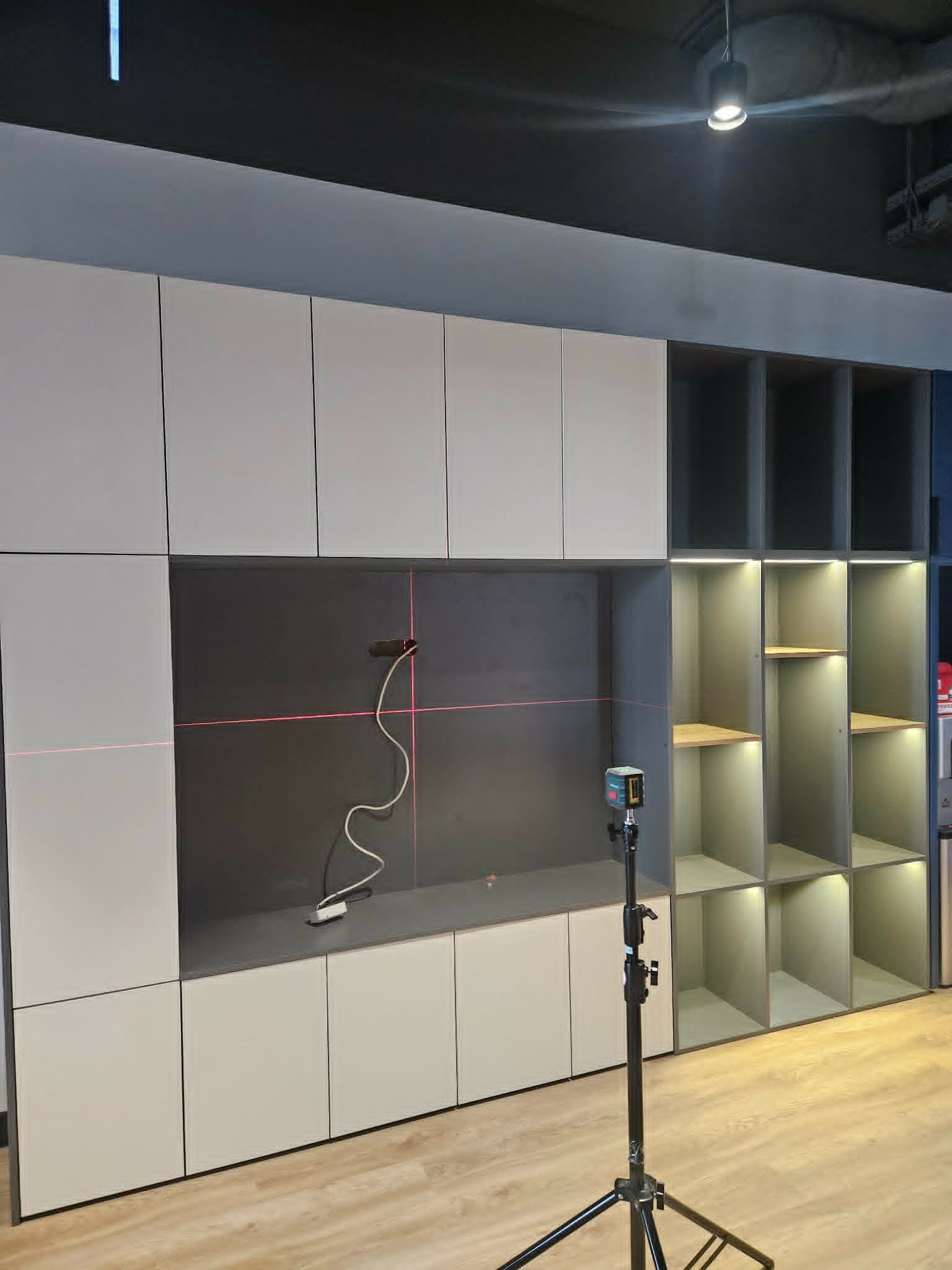 Montaż szafy wnękowej z użyciem poziomicy laserowej. Widoczne półki i zamknięte szafki w odcieniach szarości. Podłoga drewniana.