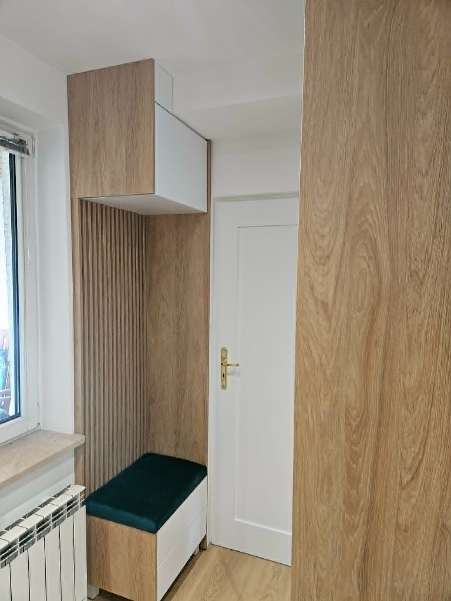 Funkcjonalna zabudowa wnęki z siedziskiem i szafką wiszącą, wykończona drewnopodobną okleiną. Obok białe drzwi i fragment grzejnika.