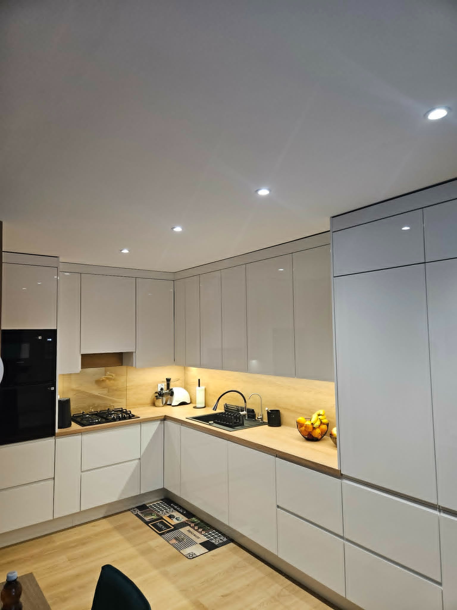 Jasna kuchnia z drewnianym blatem, oświetleniem podszafkowym i czarnym piekarnikiem. Ujęcie z perspektywy podłogi, widoczny fragment dywanika kuchennego.