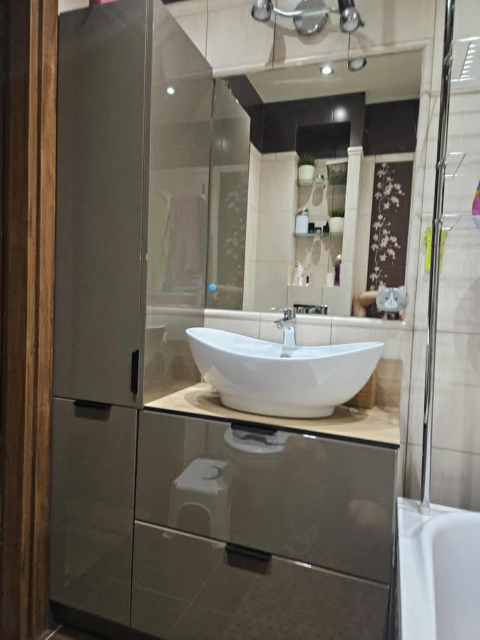 Nowoczesna łazienka z szafką pod umywalkę w kolorze taupe, białą umywalką nablatową i lustrem z oświetleniem. Widoczna część wanny i dekoracje na półkach.