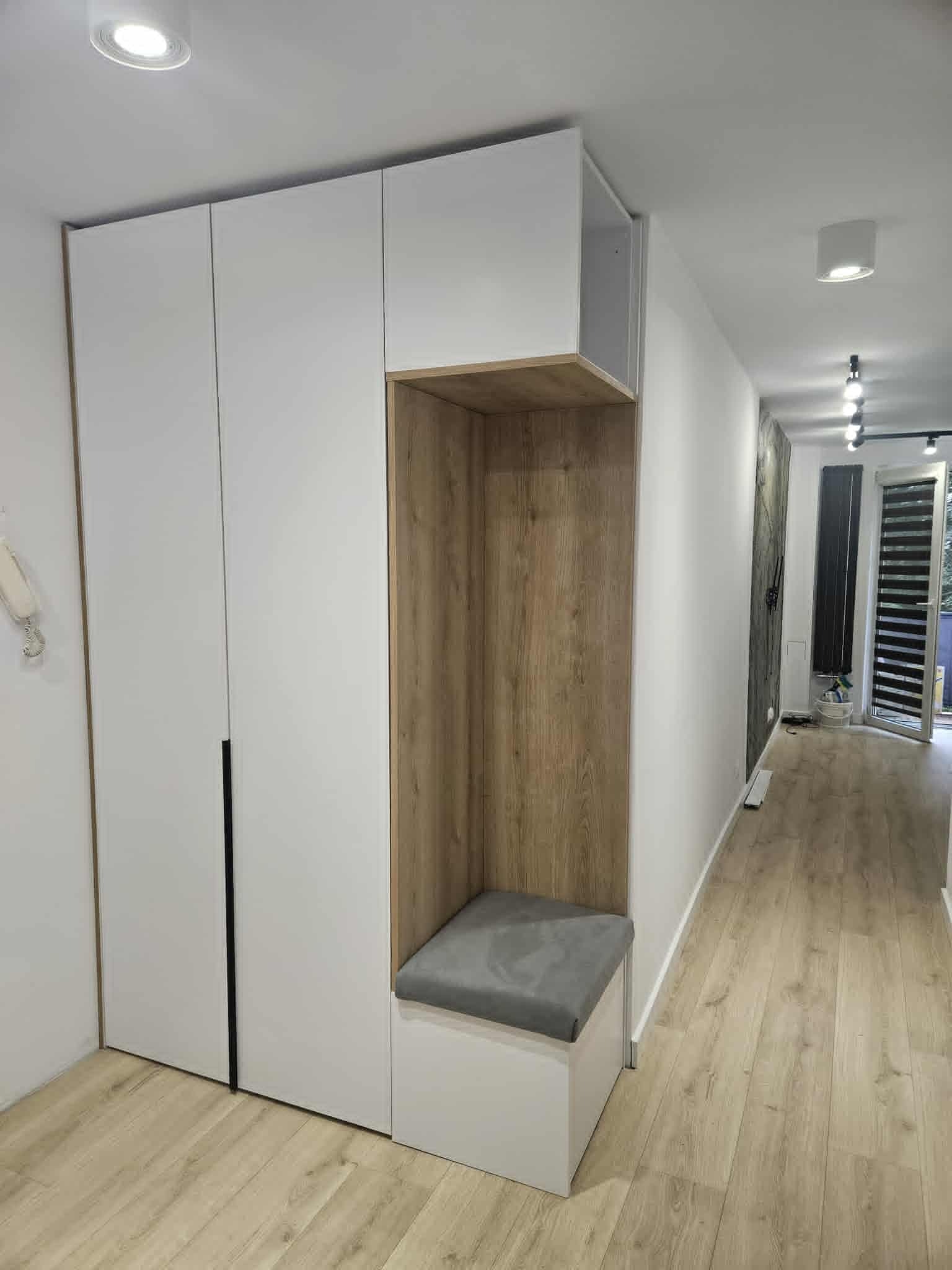 Biała szafa wnękowa z siedziskiem i drewnianą wnęką w przedpokoju. Minimalistyczny design, jasne drewno na podłodze, nowoczesne oświetlenie sufitowe.