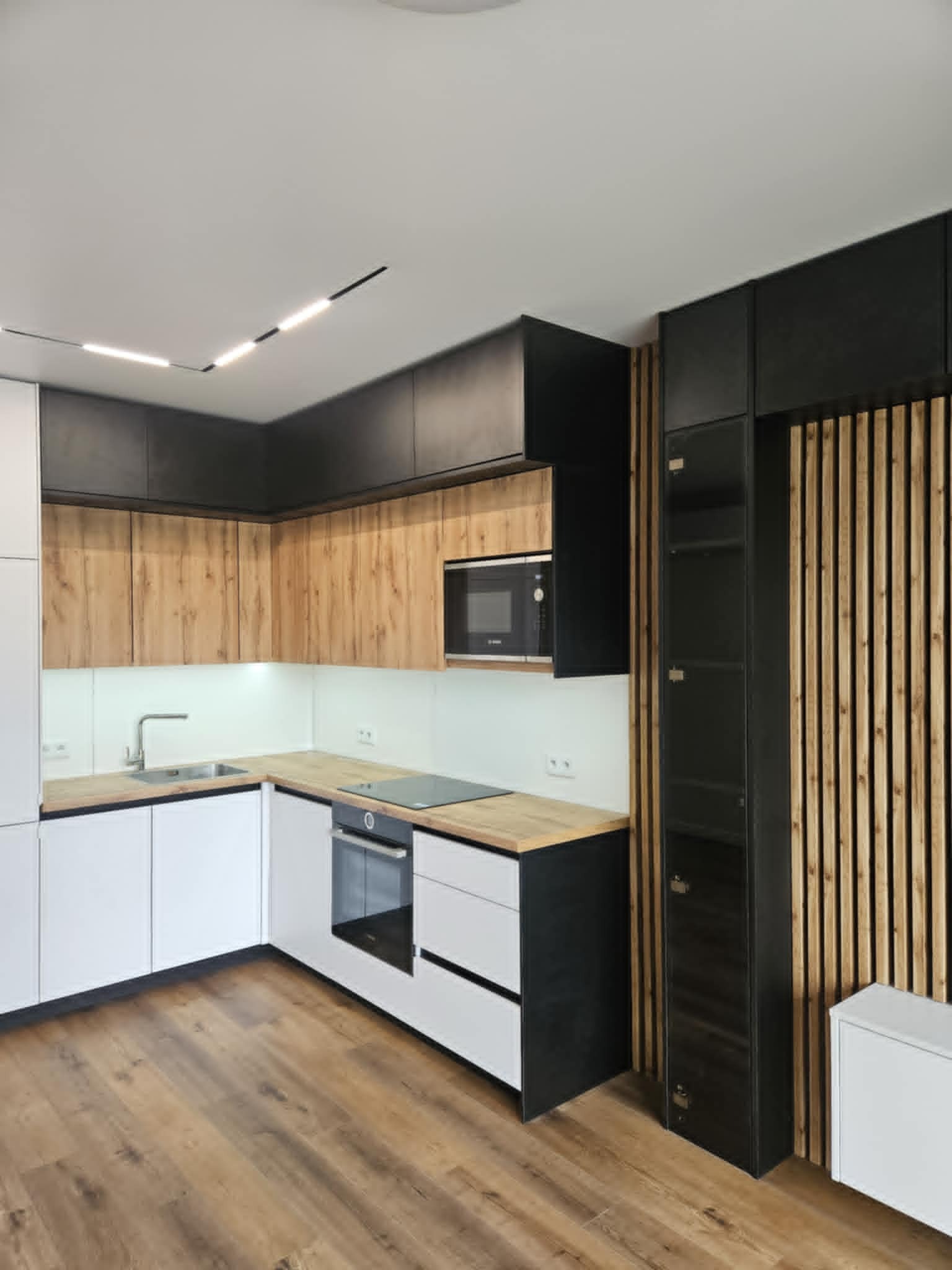 Nowoczesna kuchnia z drewnianymi frontami szafek górnych i białymi dolnymi. Czarny słupek z drewnianą ścianką działową. Oświetlenie liniowe LED.