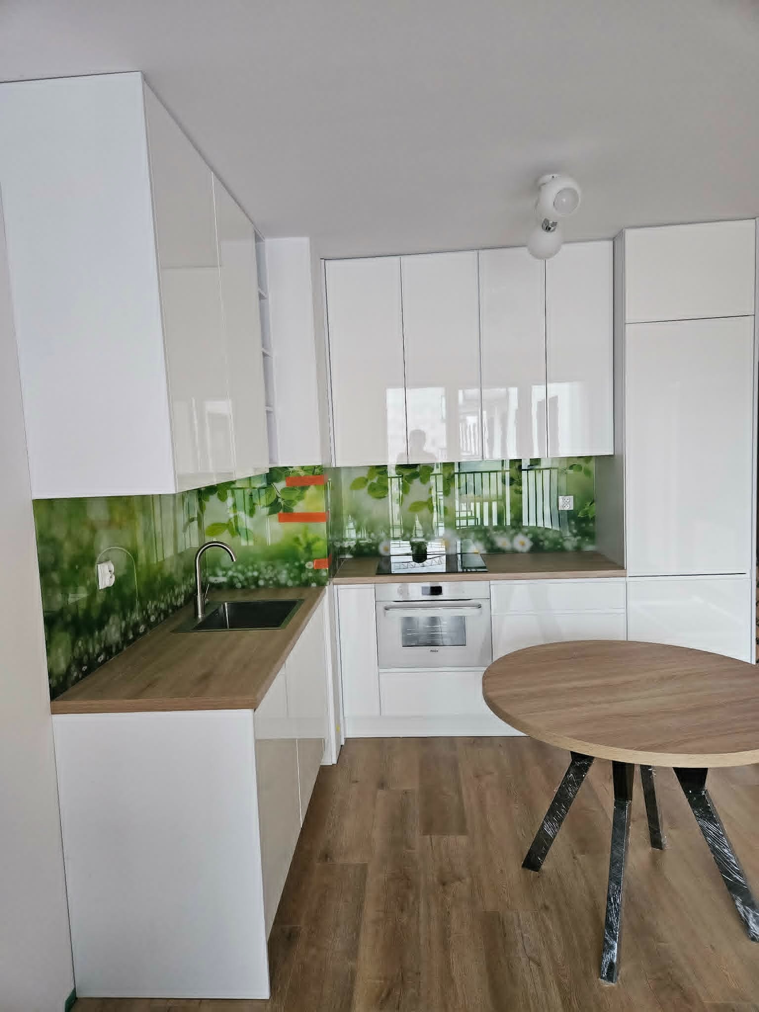 Biała kuchnia z blatem drewnopodobnym i szklanym panelem z motywem roślinnym. Okrągły stół z drewnianym blatem na metalowych nogach. Nowoczesne oświetlenie sufitowe.