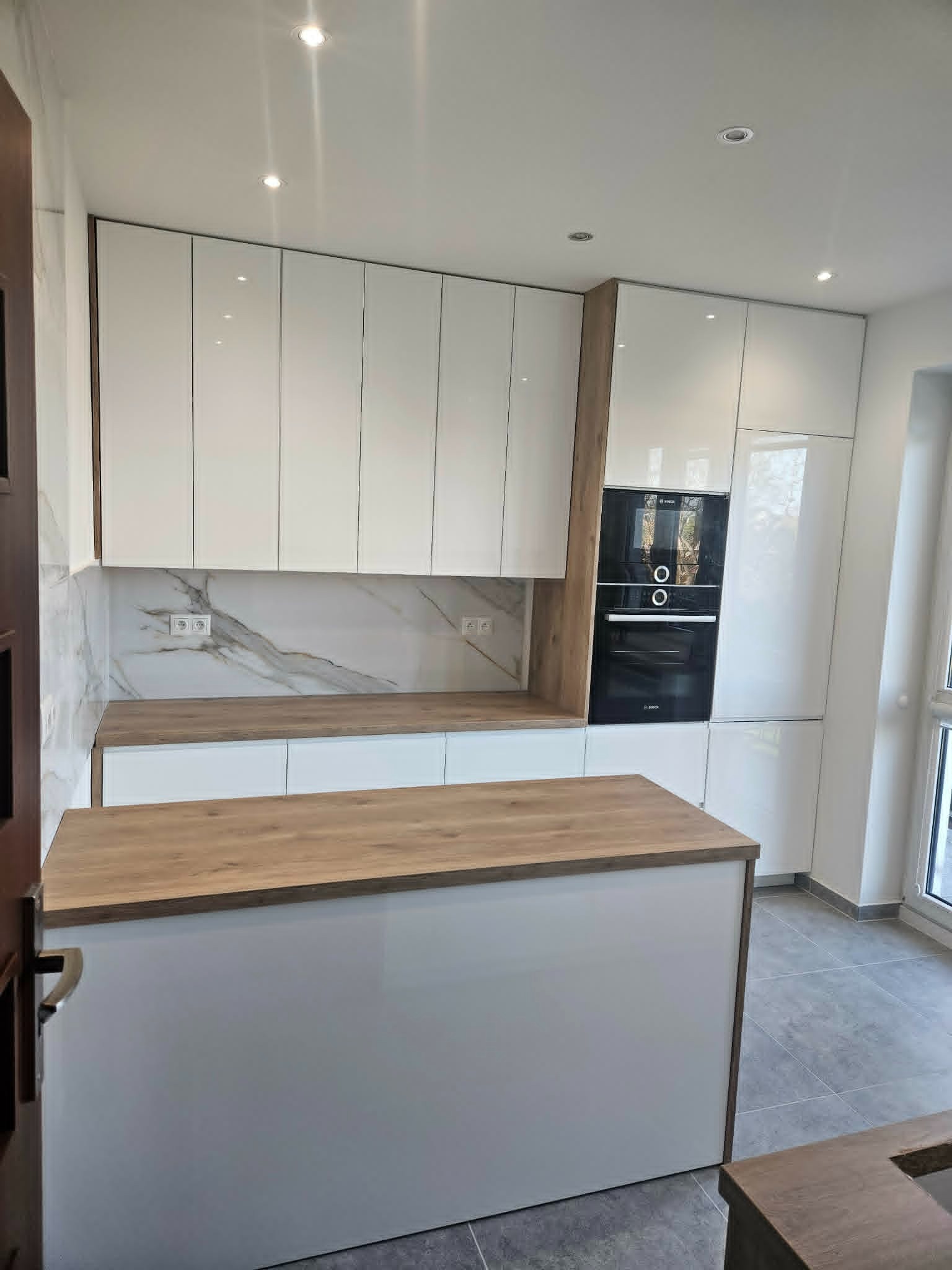 Biała kuchnia z wyspą i drewnianym blatem, fronty na wysoki połysk, marmurowy backsplash, zabudowa piekarnika, nowoczesny design, szare płytki podłogowe.