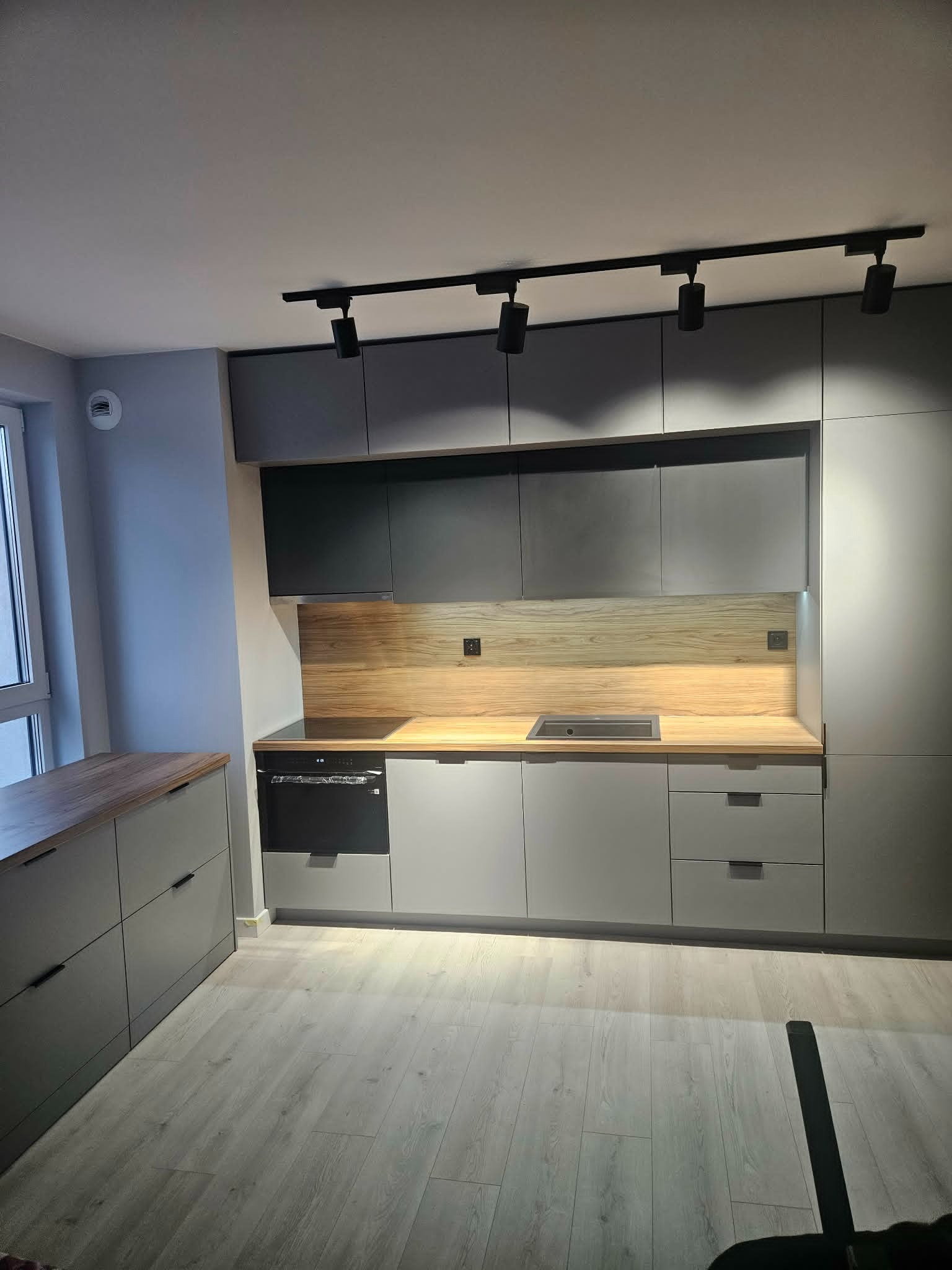 Nowoczesna kuchnia w odcieniach szarości z drewnianym blatem i oświetleniem punktowym. Minimalistyczny design, fronty bez uchwytów, zabudowa od podłogi do sufitu.