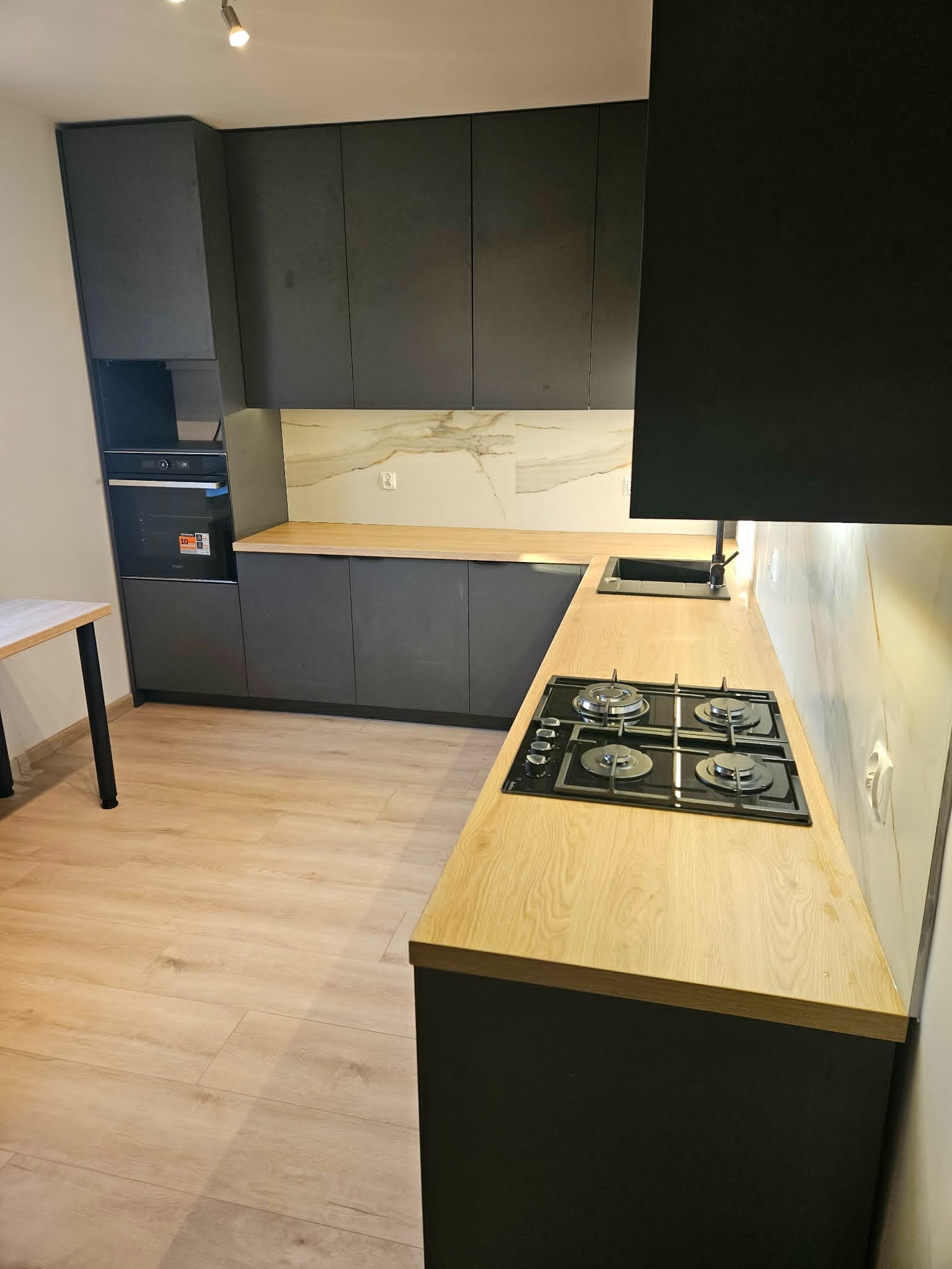 Nowoczesna kuchnia z ciemnymi szafkami i drewnianym blatem, płyta gazowa. Minimalistyczny design, jasna podłoga, marmurowe wykończenie ściany nad blatem.