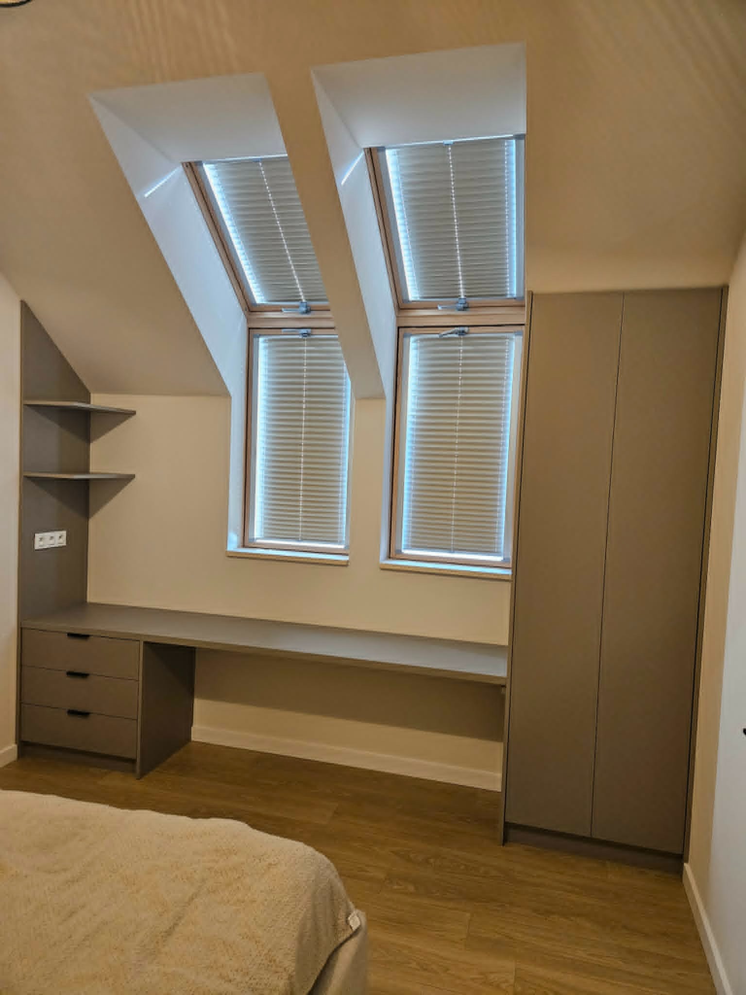 Szafa wnękowa z biurkiem i półkami w pokoju na poddaszu, szare fronty, drewniana podłoga, dwa okna dachowe z żaluzjami.