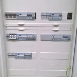 Otwarta, biała skrzynka rozdzielcza z zainstalowanymi bezpiecznikami i wyłącznikami różnicowoprądowymi, widoczny przewód ochronny.