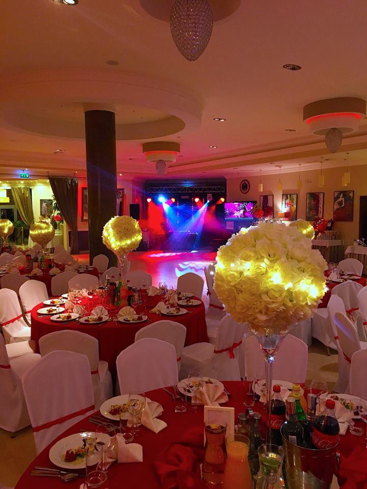 Uroczysta sala bankietowa z okrągłymi stołami nakrytymi czerwonymi obrusami, białymi krzesłami z czerwonymi wstążkami, kwiatowymi dekoracjami z podświetleniem LED w wysokich wazonach oraz sceną...