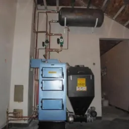 Niebieski piec na paliwo stałe z zasobnikiem, połączony miedzianymi rurami z instalacją grzewczą w Pleszewie, widoczne elementy sterujące i zbiornik wyrównawczy pod sufitem w surowym pomieszczeniu.