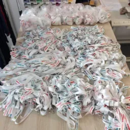 Stół roboczy zasypany stertą białych smyczy z nadrukowanymi logo firm i nazwiskami, częściowo zapakowanych w przezroczyste woreczki; w tle widoczny organizer z kolorowymi nićmi.