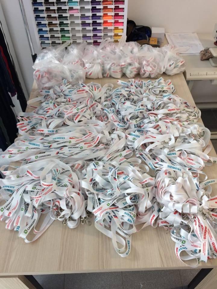 Stół roboczy zasypany stertą białych smyczy z nadrukowanymi logo firm i nazwiskami, częściowo zapakowanych w przezroczyste woreczki; w tle widoczny organizer z kolorowymi nićmi.