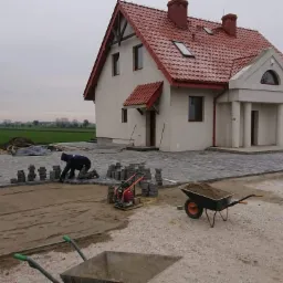 Układanie szarej kostki brukowej przed nowym domem z czerwoną dachówką; w tle widoczne pole i pochmurne niebo. Pracownik w ciemnej bluzie klęczy przy układaniu kostki, obok stoi zagęszczarka...