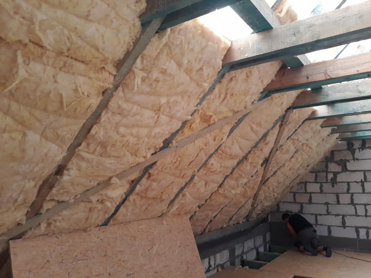 Ocieplenie poddasza wełną mineralną między krokwiami, widoczna konstrukcja drewniana dachu i fragment ściany z białego bloczka, pracownik w trakcie montażu.