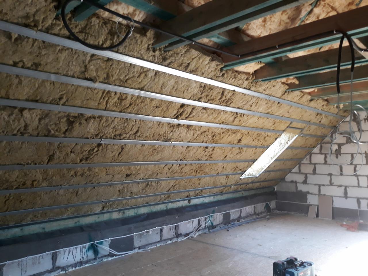 Izolacja poddasza wełną mineralną, widoczne metalowe profile konstrukcyjne i fragment muru z białych bloczków.
