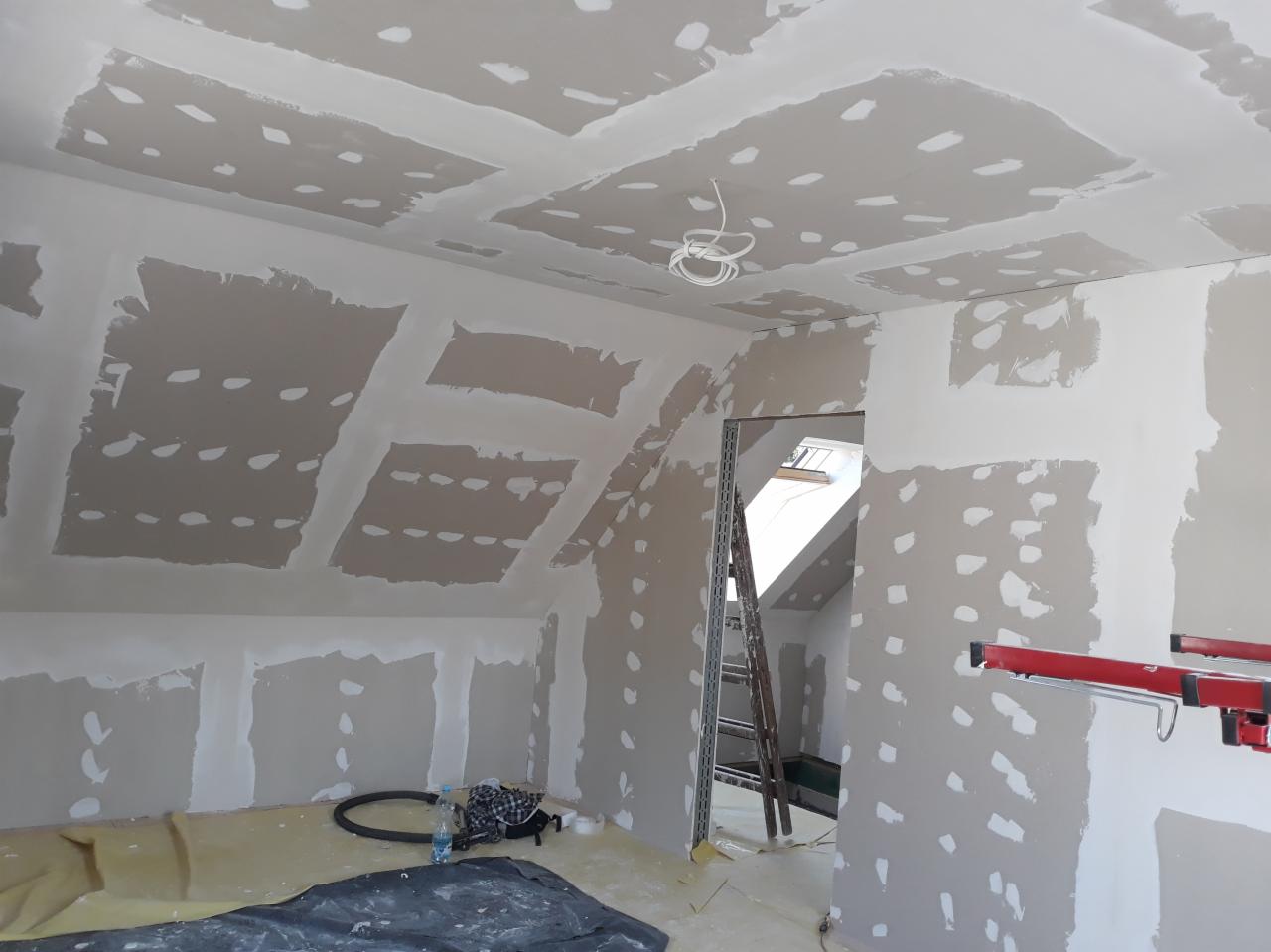 Poddasze w trakcie wykańczania: płyty karton-gips zamontowane na skośnym suficie i ścianach, wstępnie szpachlowane łączenia, widoczna drabina, przewody elektryczne i narzędzia.