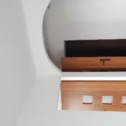 Biała ściana z zaokrąglonym narożnikiem sąsiadująca z drewnianą konstrukcją z trzema kwadratowymi otworami, ujęcie z dołu.